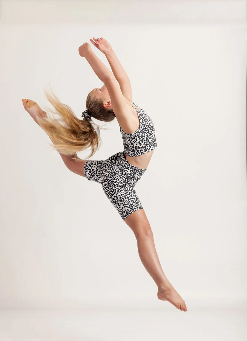 Girl in an Ignite Snow Leapord Crop top and Get moving bike shorts performing a split leap on a light background 