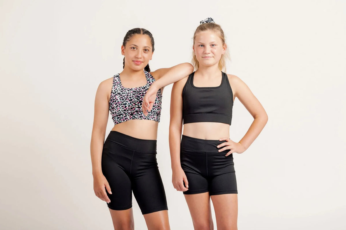Two girl, one wearing a race ready black crop and black shorts and the other a multi coloured leopard print crop and black shorts, on a light background