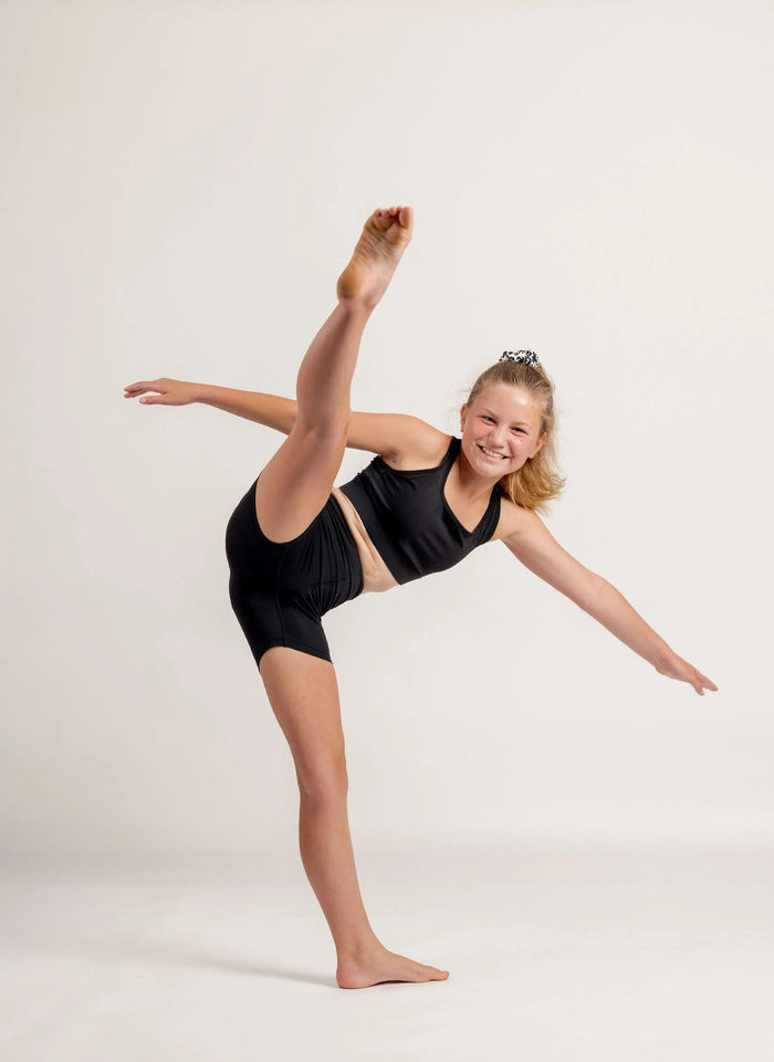 Young girl in a black crop and black mini biker shorts performing a ballet pose on a white background
