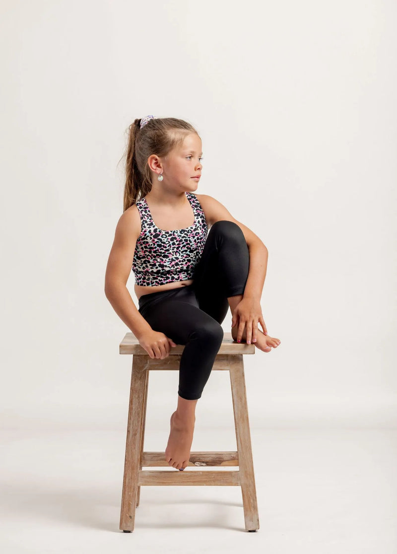 Young girl sat on a wooden stool, wearing a Multi coloured leopard print crop top and black Go Get Em 3/4 leggings on a white background