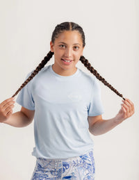 Girl wearing a Gametime light blue t-shirt with a logo, holding their braided hair against a plain background
